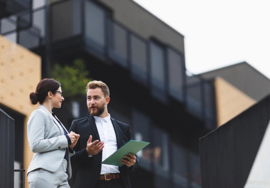 To eiendomsmeglere står og snakker sammen. I bakgrunnen, uskarpt, er en stor eiendom.